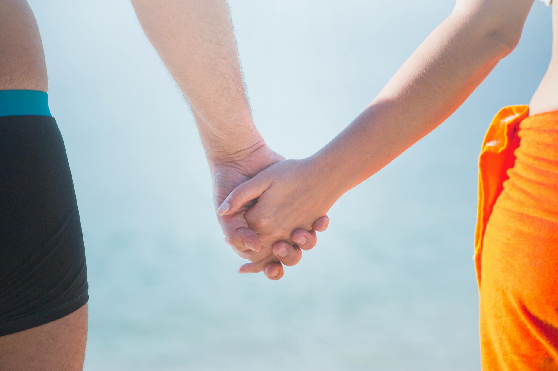 midsection of couple holding hands at beach against sky