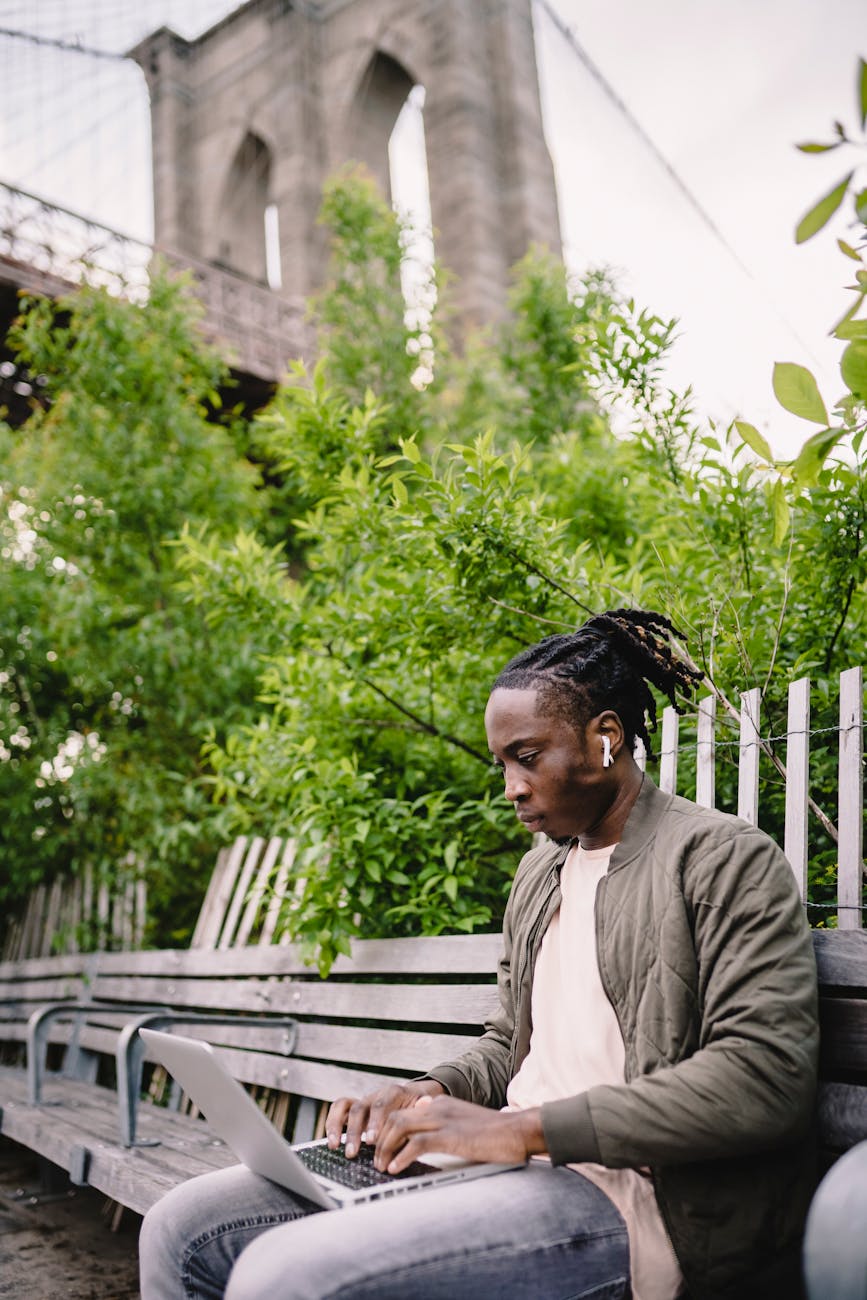 young man sitting on park bench and working on laptop