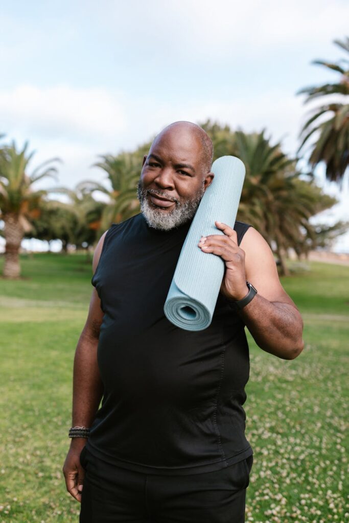 an elderly man in black tank top smiling while holding his yoga mat