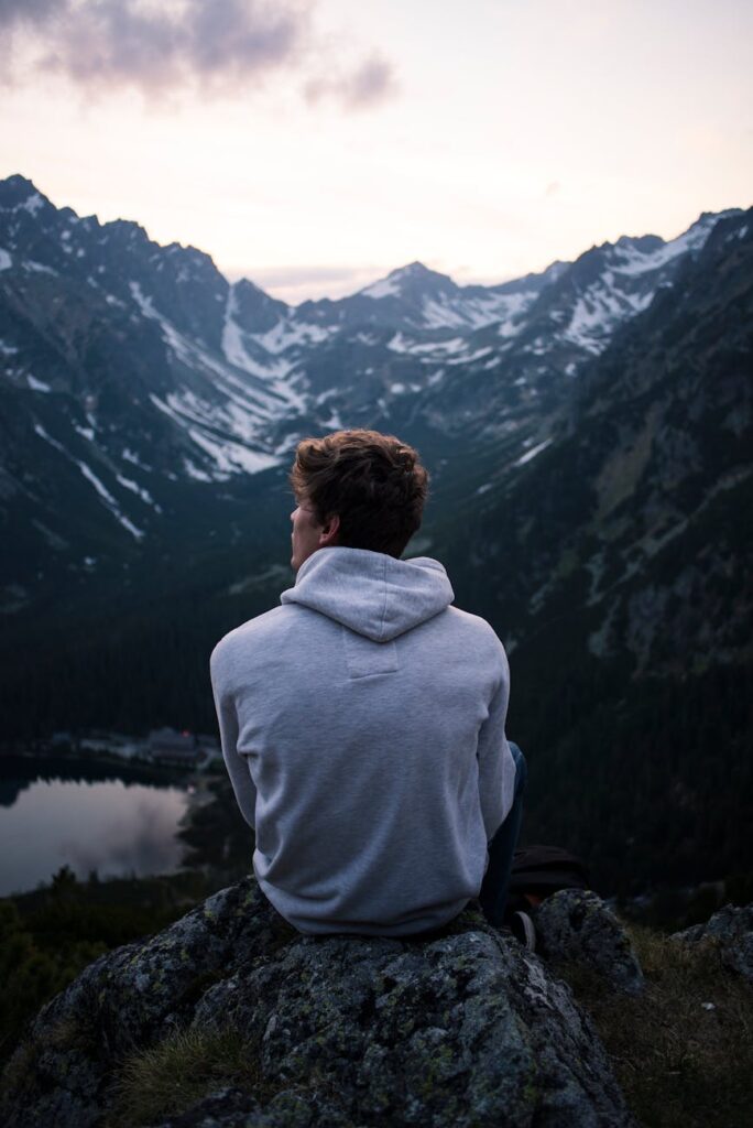 a man in white hoodie sitting on a rock across the mountains